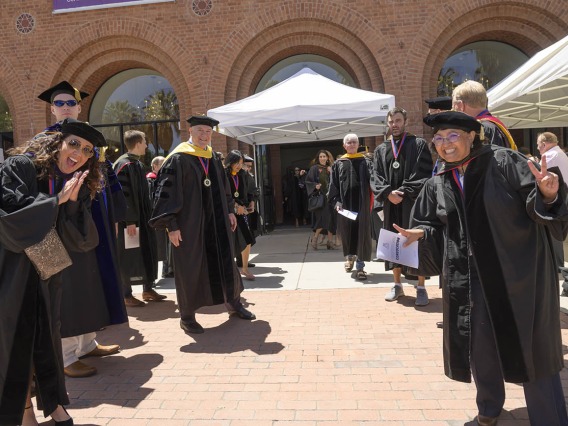 A group of Pharmacy professors stand on both sides of a walkway outside a large brick building waving and cheering. 