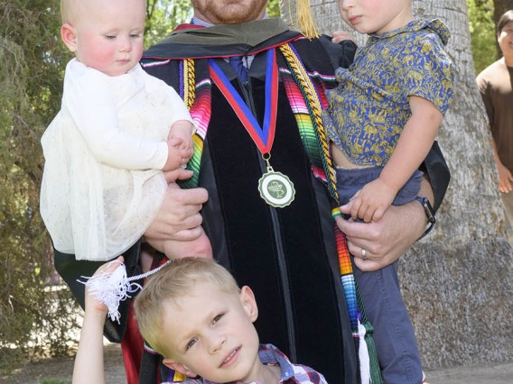 A young man with a beard wearing a graduation cap and gown holds two young children in his arms while another child stands in front of him. 