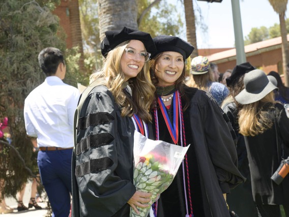 A young woman with long blonde hair in a graduation cap and gown smiles, holding flowers and posing with a female professor in graduation regalia as well. 