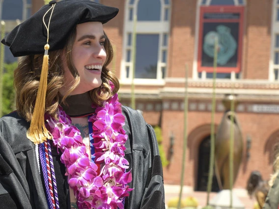 Profile of a smiling young white woman in a cap and gown with flowers around her neck with a red brick building in the background.  