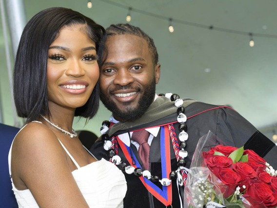 A smiling young Black man in a graduation gown holding flowers, side-hugs a young, smiling Black woman. 