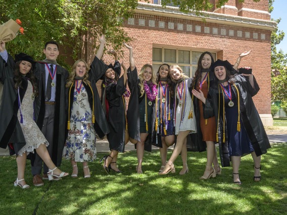 A group of nine graduates in caps and gowns stand together smiling and cheering outside. 