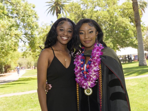 Two young Black women, one in a dress and the other in a graduation gown, side-hug and smile.