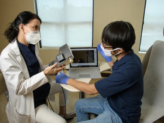 Clara Curiel Lewandrowski, MD, talks with Dongkyun Kang, PhD, as she holds the portable confocal microscope the two are developing to provide advanced skin cancer diagnostic options in the clinic. The portable device will help doctors diagnose and excise skin cancer while reducing the wait for biopsy results.