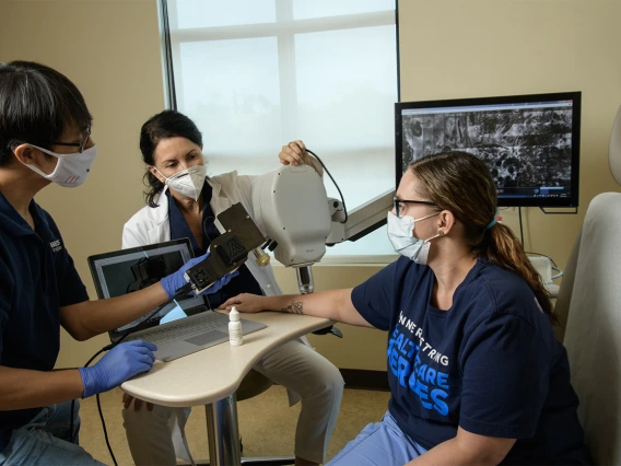 Clara Curiel Lewandrowski, MD, and Dongkyun Kang, PhD, prepare to do a side-by-side test of the confocal microscope and the portable unit to compare the image quality. Dr. Curiel is aligning the larger device, while Dr. Kang prepares to aim the handheld device at a nearby spot on Savannah Barber’s arm. Barber is a medical assistant at Banner – University Medical Center.