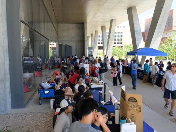 A crowd of people mill about information tables in an outside patio next to a building. 