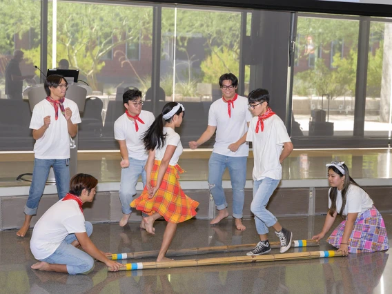 Seven students from the UArizona Filipino American Student Association perform a dance with two long wooden poles on the ground that they hop over.  