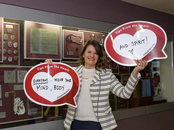 White woman with brown hair and and striped jacket holds up two heart posters in front of a display cabinet