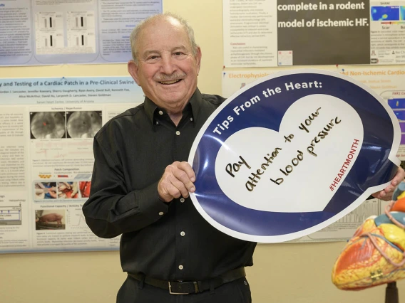 Older, balding man in black shirt holds a heart poster that says Pay attention to your blood pressure. 