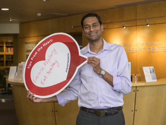 Man with brown skin and short dark hair wearing a button-up shirt holds a heart sign. 