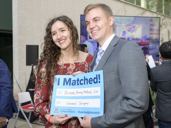 A young adult, light skinned man and woman stand together smiling and holding a Match Day poster.