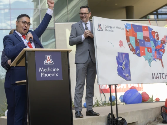 A young adult male wearing a suite and tie raises a hand in celebration while standing at a podium. A map of the US is in the background. 