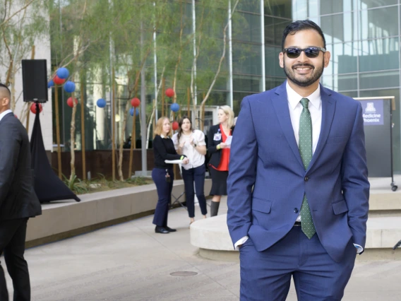 A young adult man in a suite and tie with short dark hair, sun glasses and a beard smiles in an outdoor setting. 