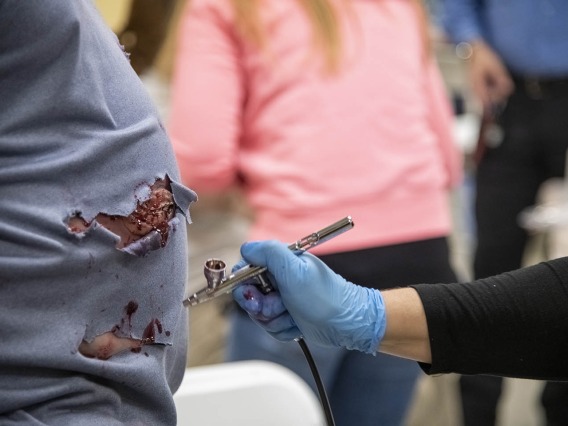 Deana Ann Smith, BS, BSN, RN, healthcare simulation educator for ASTEC, air brushes blood-red special-effects makeup on a man’s stomach for an emergency exercise. 