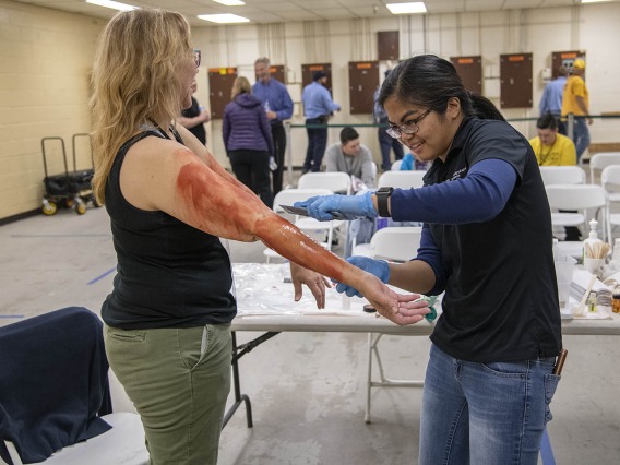 With outstretched arms, volunteer Pamela Leonard prepares to roleplay an airplane passenger injured by a midair explosion. Merryl Lopido, simulation operations specialist for ASTEC, uses special-effects makeup to create fake burns.