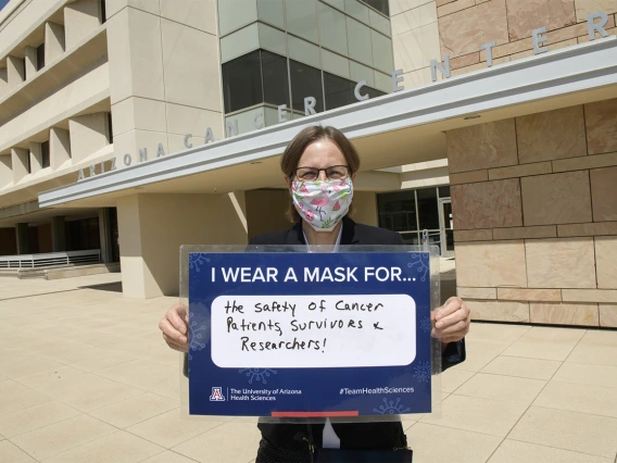 UArizona Cancer Center director Joann Sweasy, PhD, holds her sign in front of the Cancer Center’s Levy and Salmon buildings  in Tucson. She wears a mask for “the safety of cancer patients, survivors and researchers.”