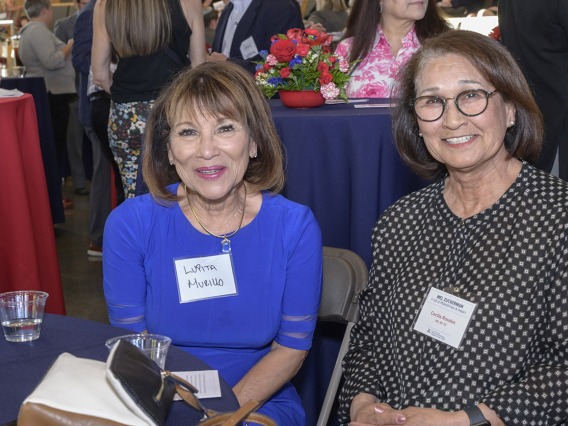 Two older women with shoulder-length brown hair sit at a table smiling.