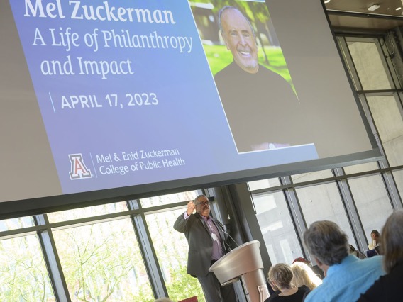 A tall dark-skinned man in a suite stands at a podium jesturing as he speaks under a large video display of Mel Zuckerman.