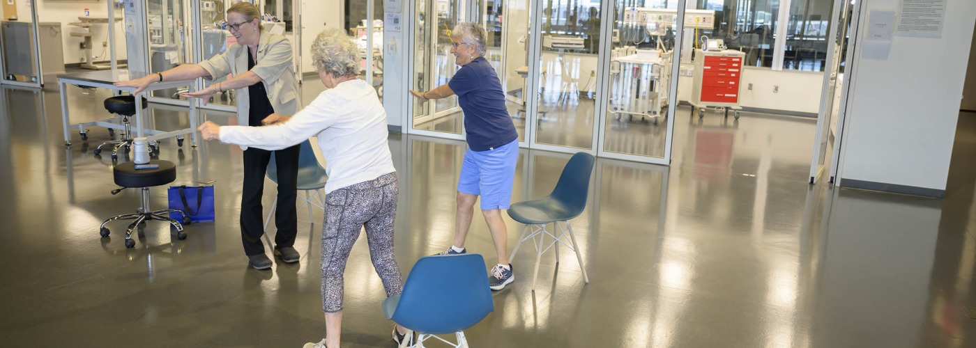 two elderly women stretch their arms out while following the standing movements of a female physical therapist