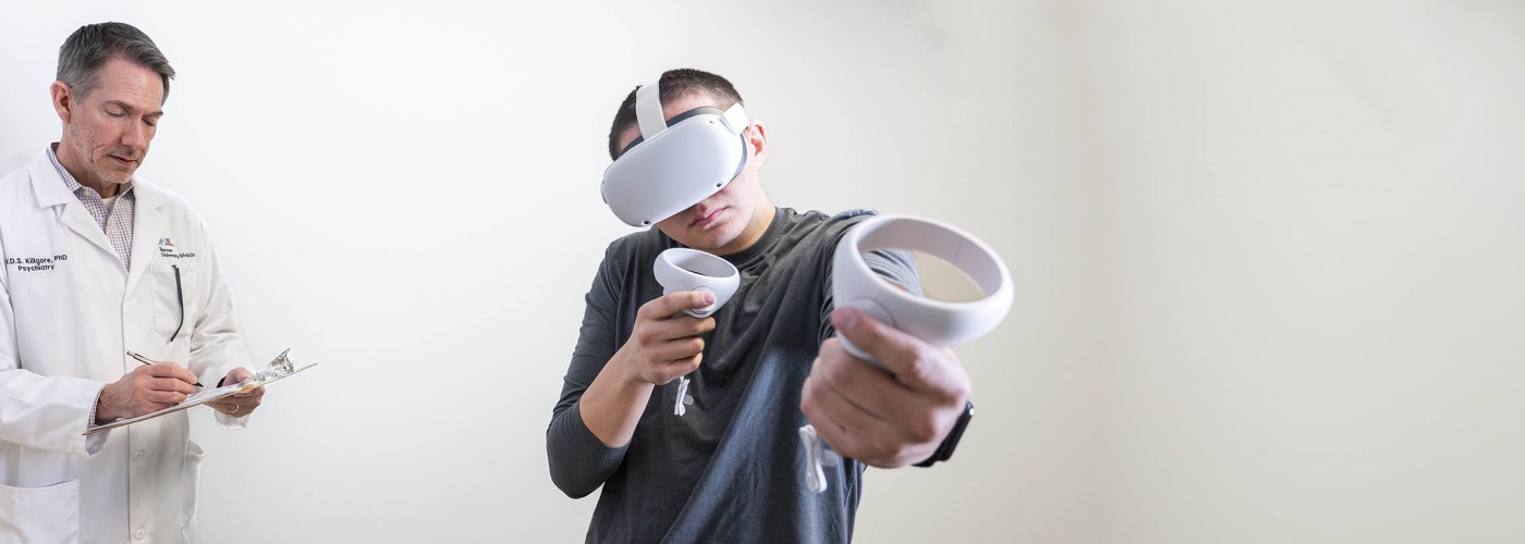 research holding clipboard stands behind male study participant wearing VR goggles and holding VR paddles
