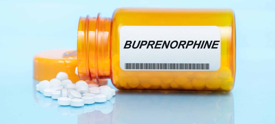 medication bottle labeled buprenorphine is laying on its side with white pills spilling out onto a light blue table