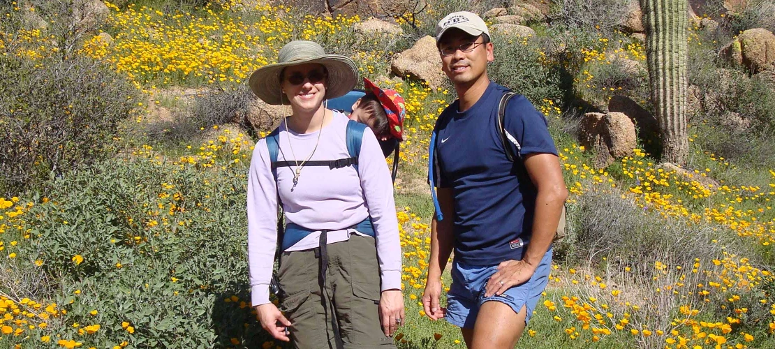 A couple with a baby in a carrier pose for a photo while hiking in the Sonoran desert