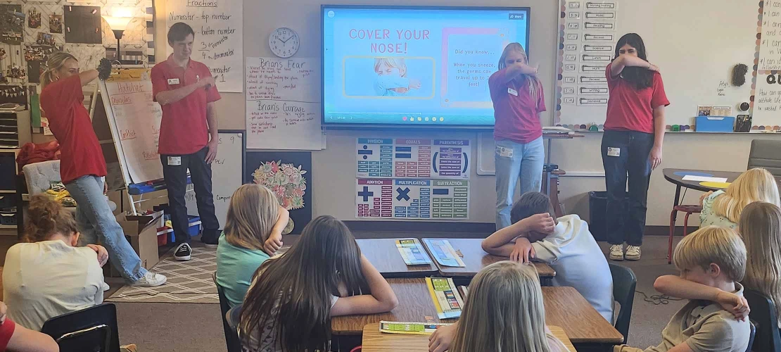 College students in red shirts stand in front of an elementary school class and demonstrate how to cover your nose as part of a health literacy presentation