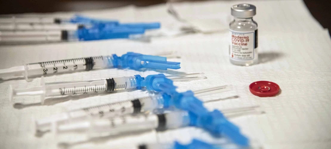 A table covered by a while cloth holding a vial with label “Moderna COVID-19 vaccine” and several syringes with needles ready for vaccination injections