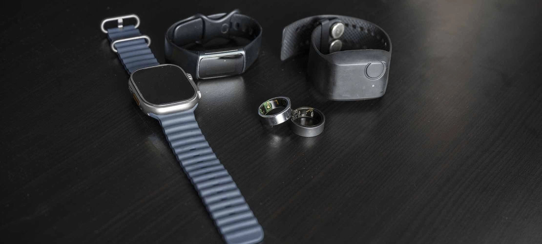 Apple Watch, Fitbit, Garmin watch, and two smart rings on a dark wood table