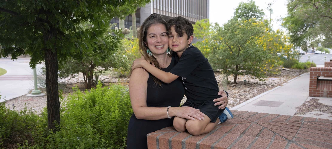 A young boy kneels on a raised surface and reaches out to hug his mother