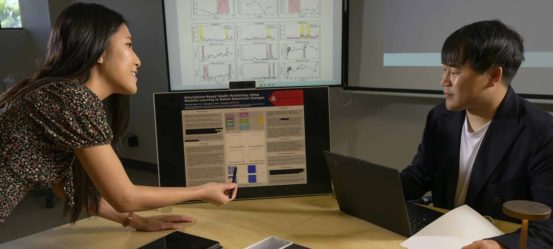 A graduate student, pointing to a small digital monitor, explains a research poster to a faculty mentor