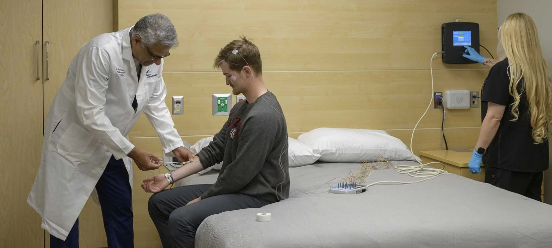 A doctor applies sensors to the arm of a person sitting on a bed in a sleep lab while another person monitors the sensor output on a small digital device on the wall.