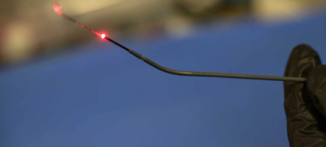 A close-up of a gloved hand holding a thin, flexible falloposcope with a red light emitting from the tip, set against a blurred lab background.