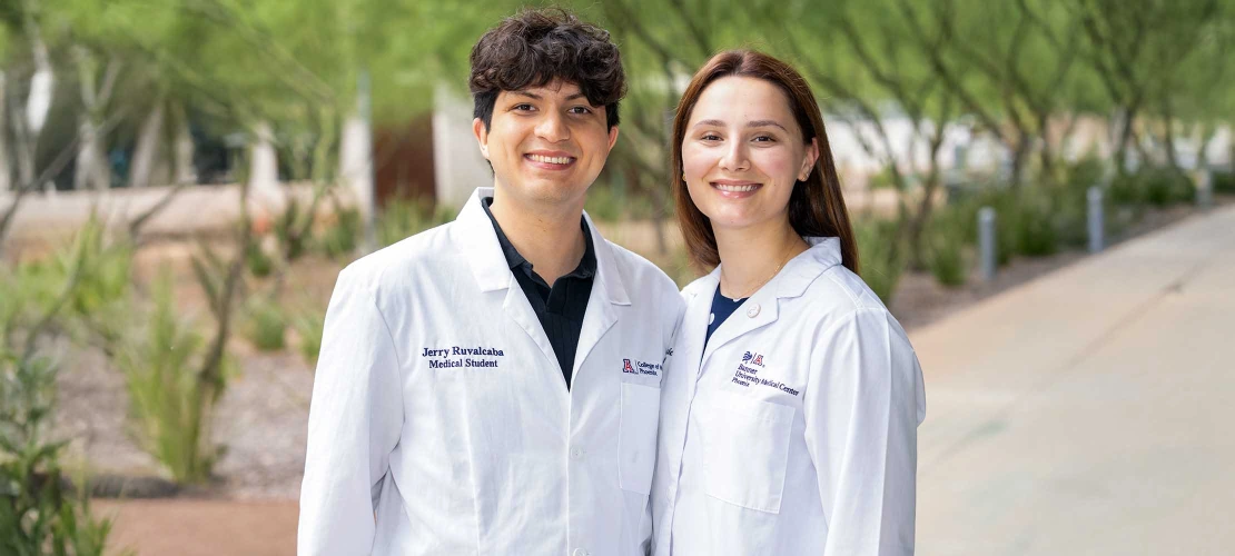 Medical students Jerry Ruvalcaba and Katie Simic stand side-by-side outdoors, smiling in their white lab coats embroidered with titles from the University of Arizona College of Medicine - Phoenix.