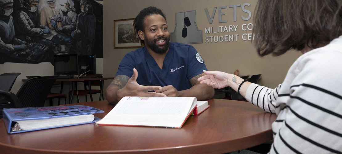 MEPN student Tery’ance Horace sits at a table with textbooks in front of him talking to a woman in front of a VETS Military Connected Student Center sign.