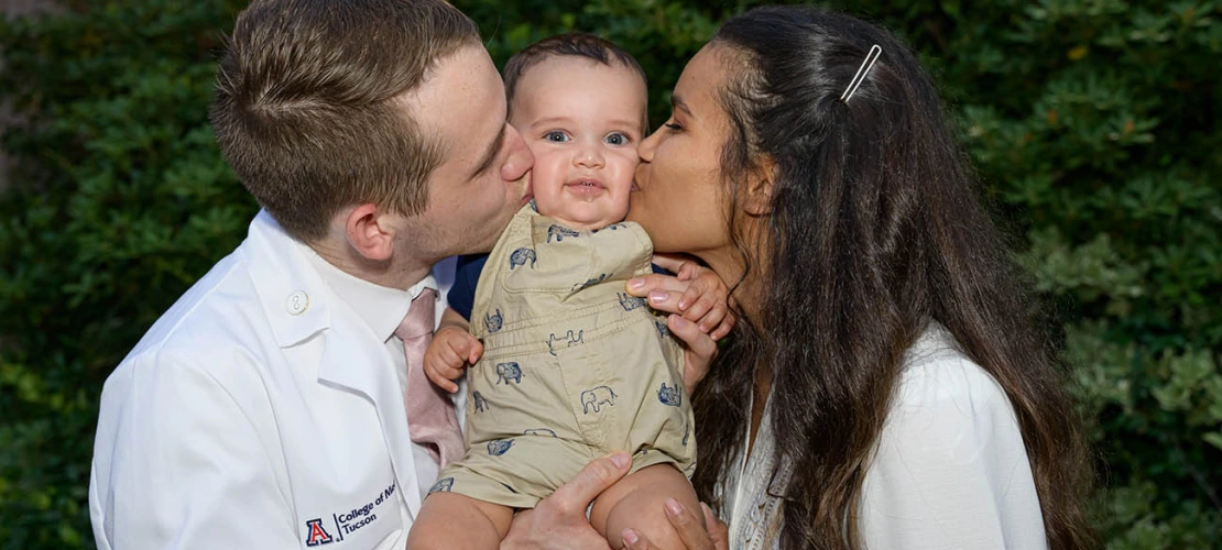 Spencer Holmes and his wife, Aiysha Holmes, celebrate his new white coat by kissing their baby, Eli, after the UArizona College of Medicine – Tucson Class of 2026 white coat ceremony.