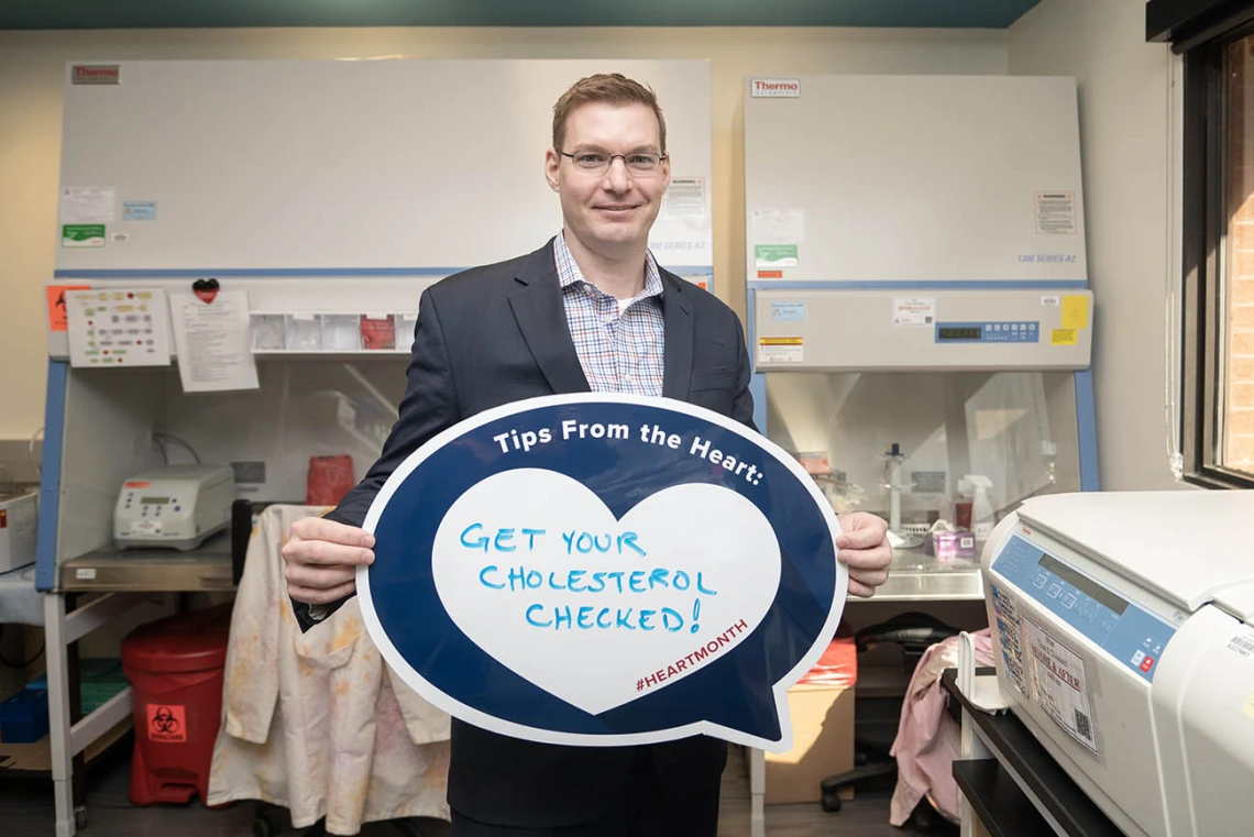 Tall, white man in dark blue sport jacket stands in a lab holding a heart poster. 