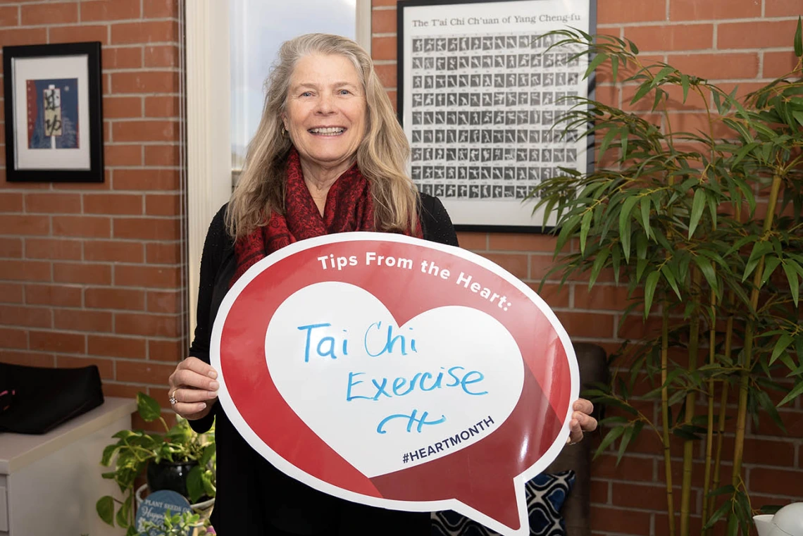 Middle-aged white woman standing in front of brick wall and plant holding heart poster.