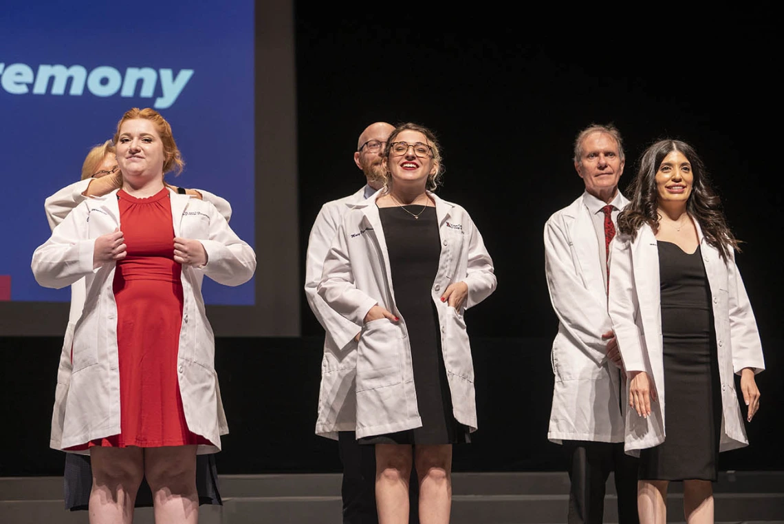 Three female Pharmacy students are presented their white coats by faculty members standing behind them on a stage. 