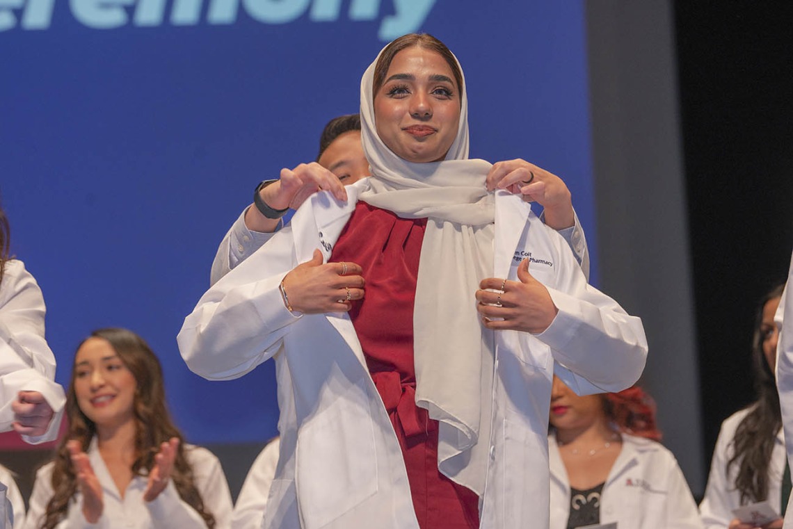 A young woman in a head scarf is presented her white coat by a faculty member standing behind her. 