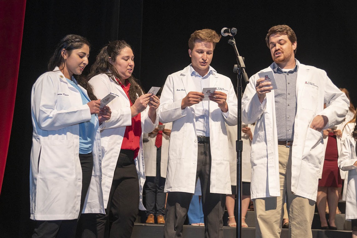Two young women and two young men in white pharmacy coats stand in front of a microphone reading from cards they are holding. 