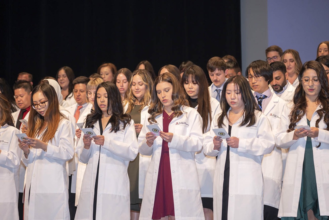 Dozens of pharmacy students wearing white coats stand on a stage reading from cards in their hands. 
