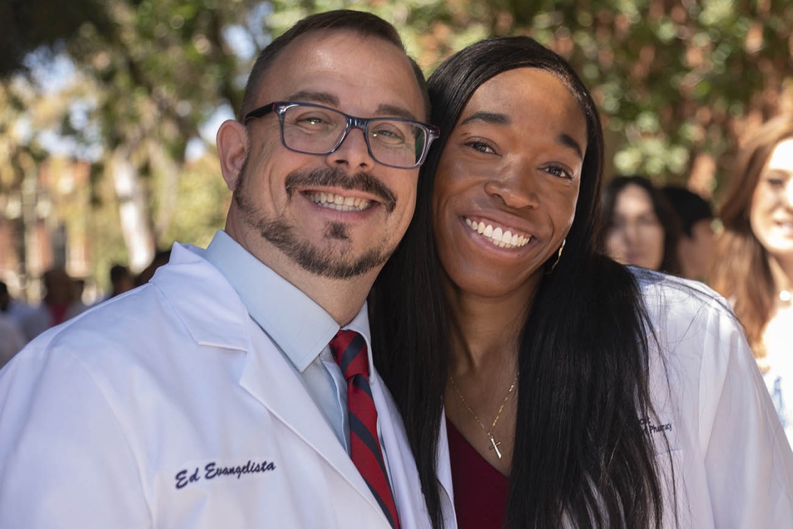 A young man and woman in pharmacy white coats smile and side-hug. 