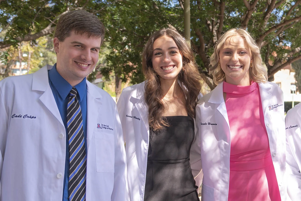 Three pharmacy students wearing white coats, a male and two females, stand next to each other smiling. 