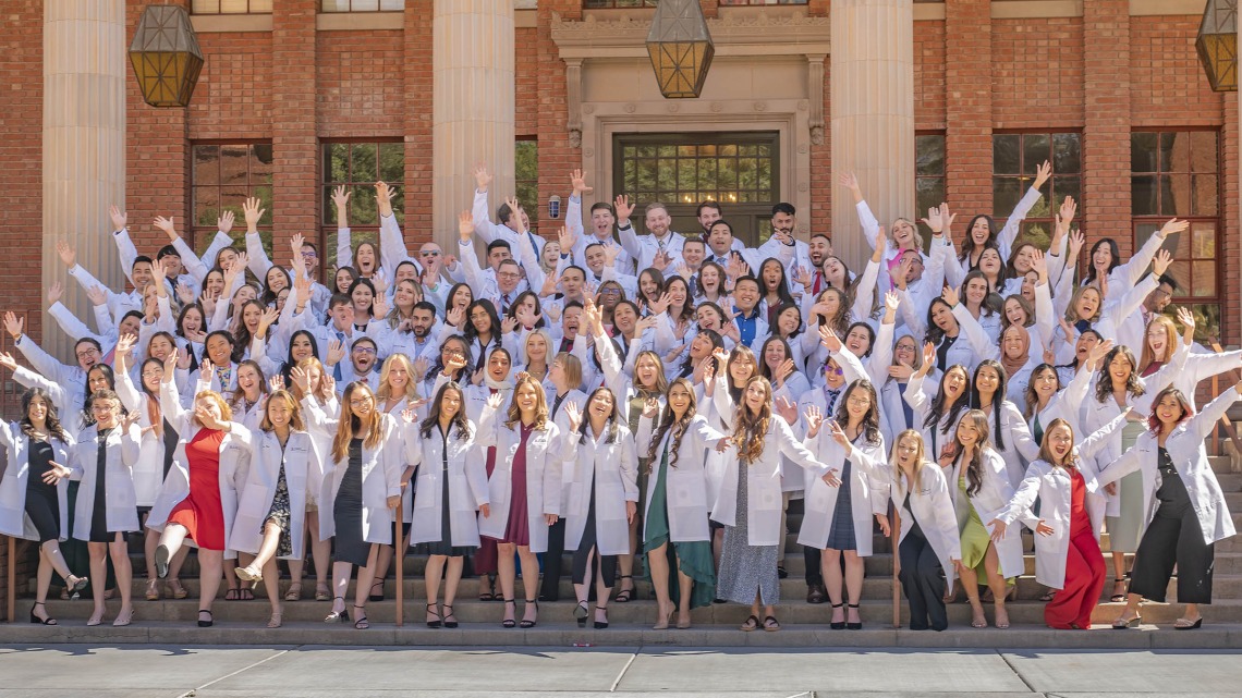 Large group of Pharmacy students wearing white coats wave their hands in celebnration.