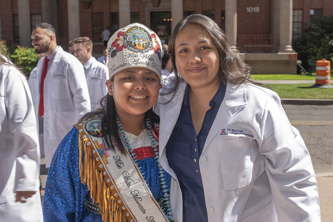 A young woman wearing traditional Native American clothing and beeded crown stands with a woman in a white pharmacy coat smiling. 