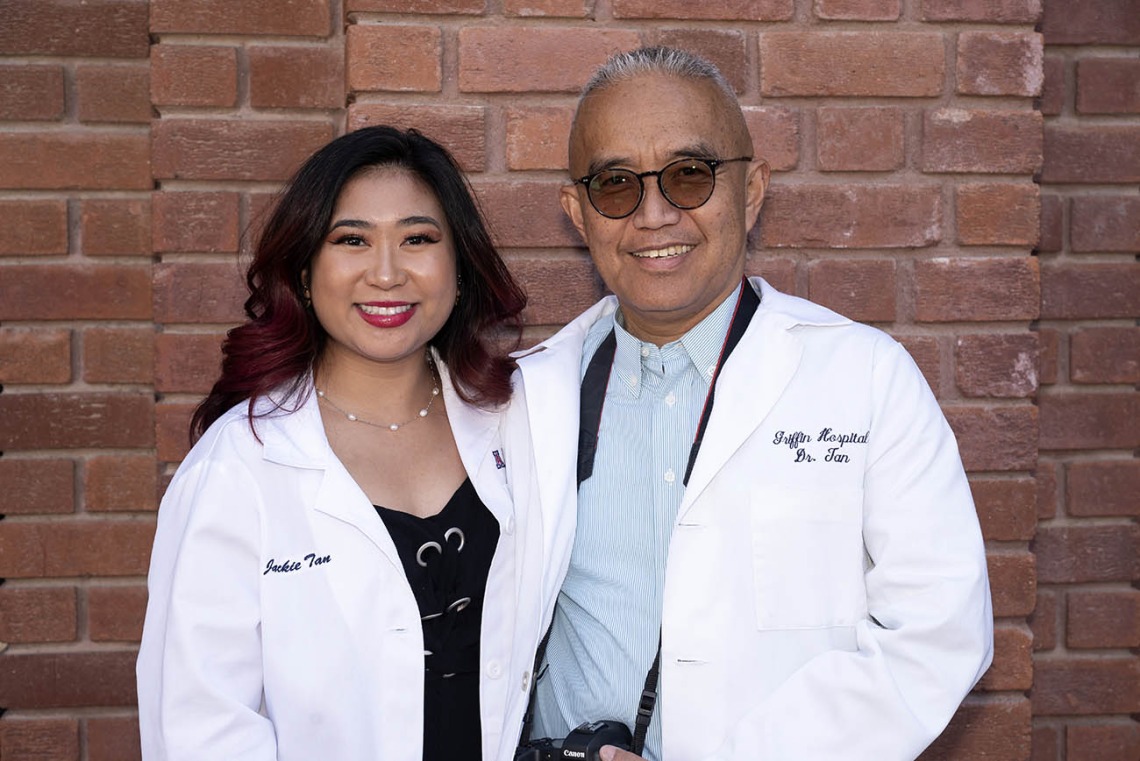 A young Asian woman in a pharmacy white coat stands with her father in a medical white coat, both smiling. 