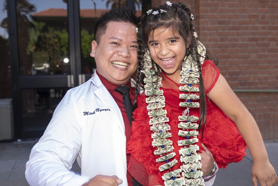 An Asian man in a pharmacy white coat holds a young girl in a red dress wearing a necklace of dollar bills. Both are smiling. 