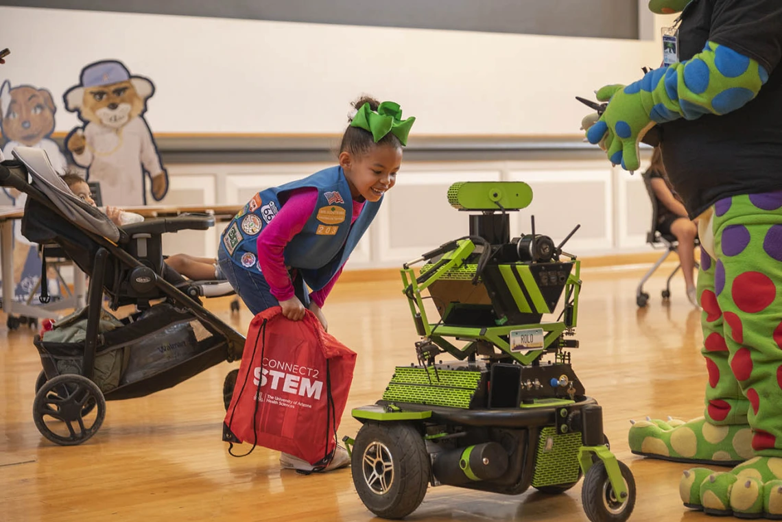 A young girl scout leans over looking closely at a green robot. 