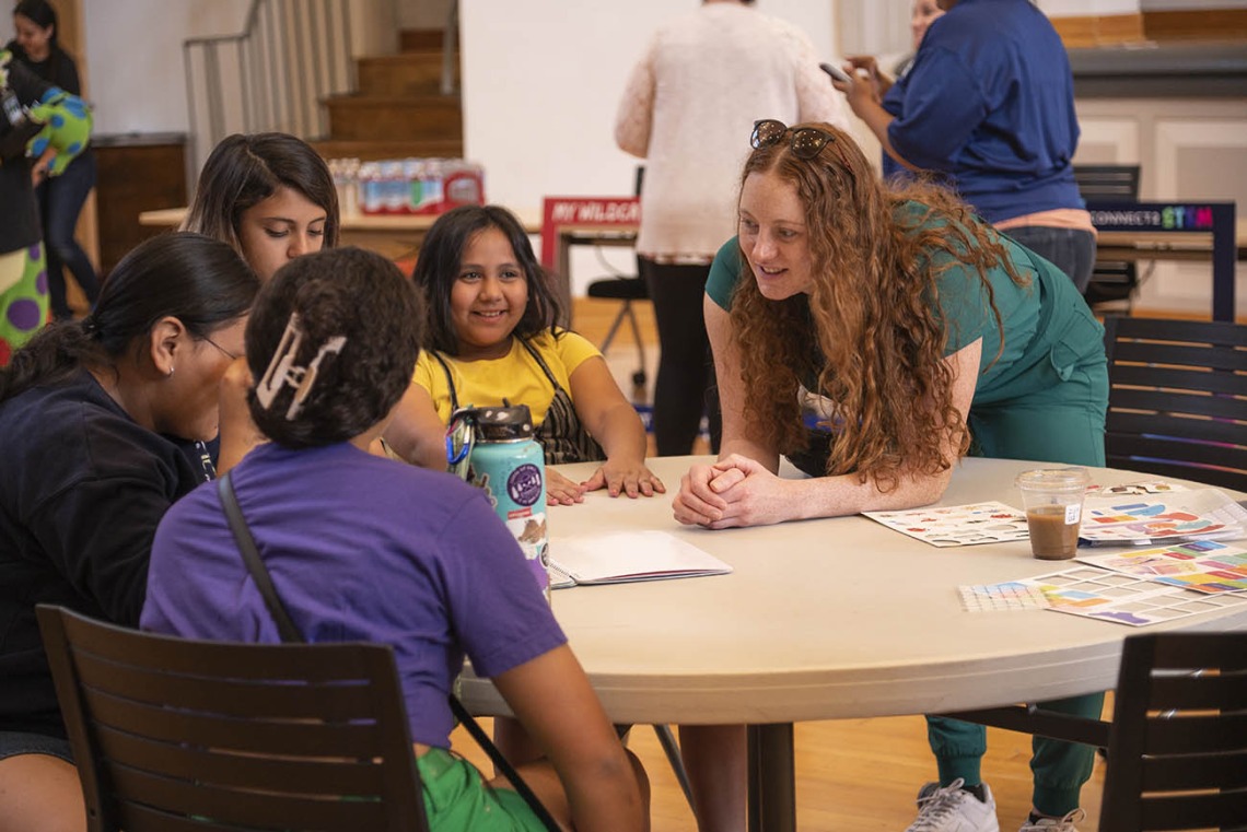 A young female medical student leans over a round table talking with a group of girl scouts.